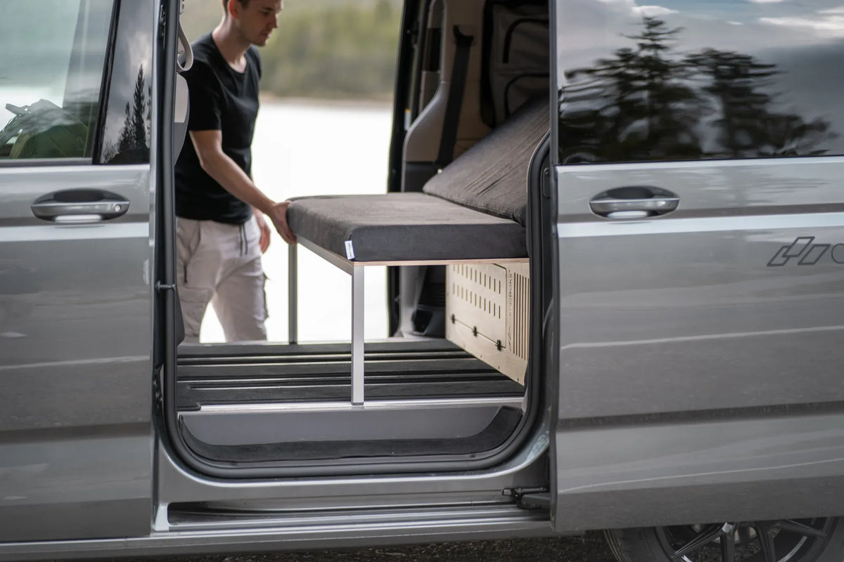 Ein Mann baut das CampBoks-Bett durch die offene Schiebetür des Vans auf.