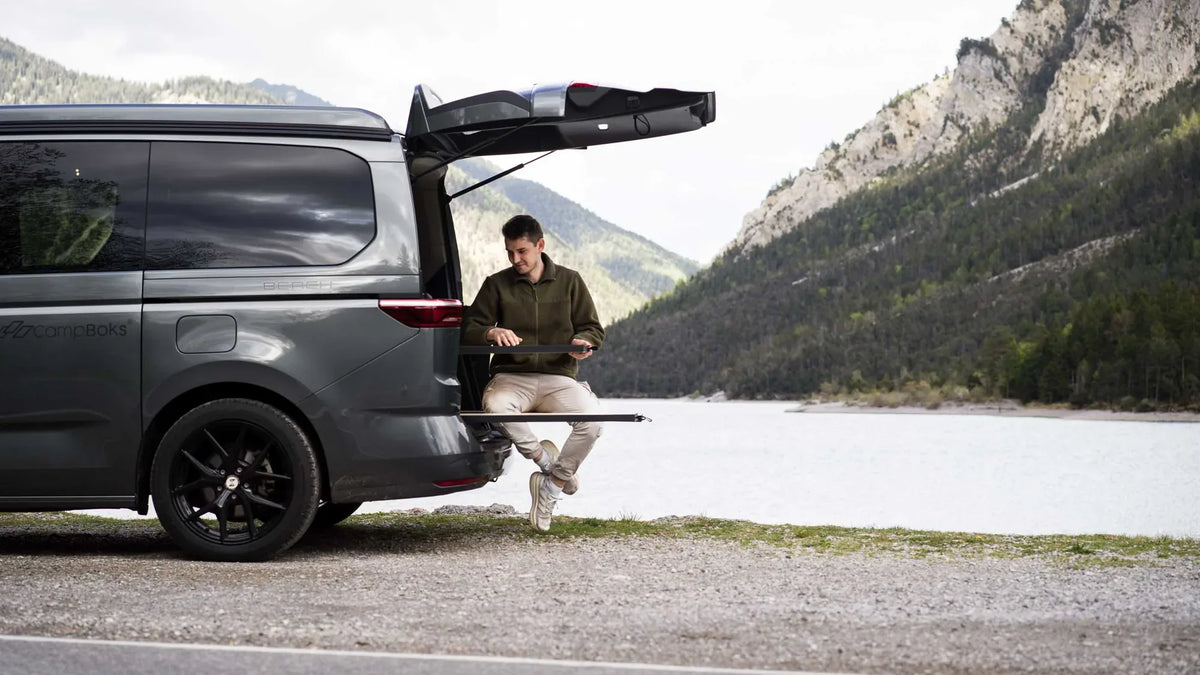 Ein Mann sitzt auf der CampBoks-Sitzbank am Fahrzeugheck vor einem See und hohen Bergen.